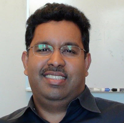 Jayachandran Kizhakkedathu, a South Asian researcher stands in front of a white board. He is wearing gold rimmed glasses and a dark burgundy shirt.